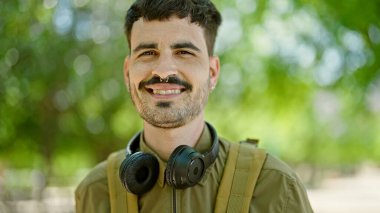 Sırt çantalı ve kulaklıklı genç İspanyol turist parkta gülümsüyor.