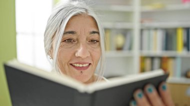 Kütüphanede gülümseyen, orta yaşlı, kır saçlı, kitap okuyan bir kadın öğrenci.