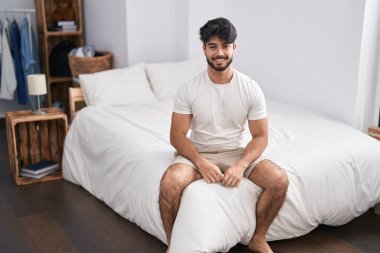 Young hispanic man smiling confident sitting on bed at bedroom