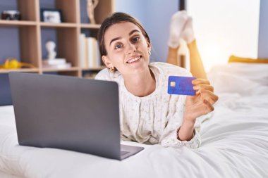 Young caucasian woman using laptop and credit card sitting on bed at bedroom