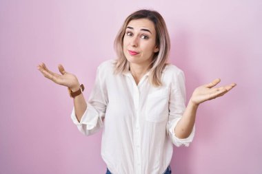 Pembe arka planda duran, elleri ve kolları havada, kafası karışmış genç ve güzel bir kadın. şüphe kavramı. 