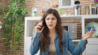Genç, güzel, İspanyol bir kadın yemek odasında elinde kızgın bir ifadeyle elinde kredi kartıyla akıllı telefondan konuşuyor.