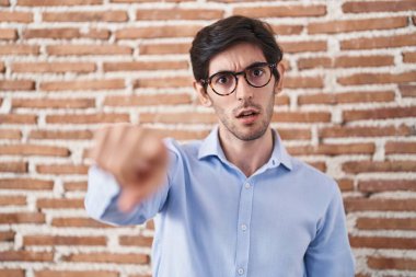 Young hispanic man standing over brick wall background pointing displeased and frustrated to the camera, angry and furious with you 