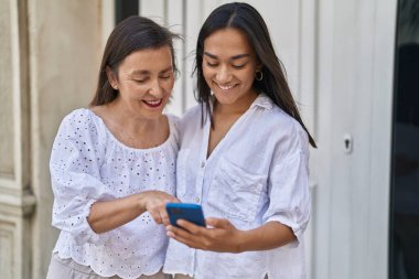 Two women mother and daughter smiling confident using smartphone at street