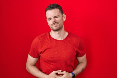 Kırmızı arka planda duran genç İspanyol adam midesi üstünde duruyor çünkü hazımsızlık, acı verici bir hastalık kendini iyi hissetmiyor. Ağrı kavramı. 