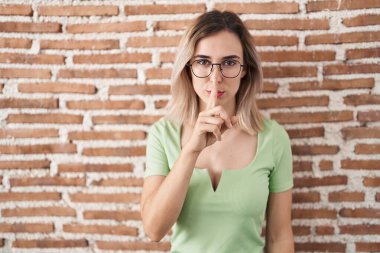 Genç ve güzel bir kadın tuğla duvarın üzerinde duruyor ve parmakları dudaklarında sessiz olmak istiyor. sessizlik ve gizli konsept. 