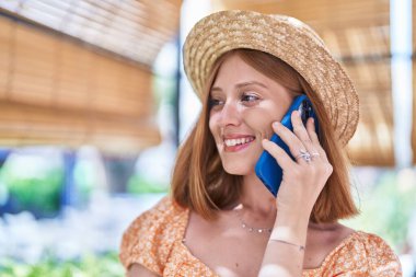 Young redhead woman tourist wearing summer hat talking on smartphone at street