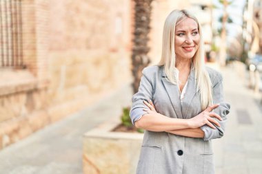 Genç sarışın kadın gülümsüyor. Kollarını kavuşturup caddede duruyor.