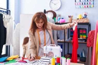 Middle age woman tailor holding t shirt writing on notebook at clothing factory