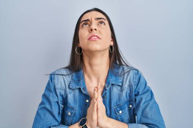 Mavi arka planda dikilen İspanyol kadın elleri bağlı yalvarıyor ve dua ediyor. Yüzünde umut ifadesi var. Çok duygusal ve endişeli. Yalvarıyor. 