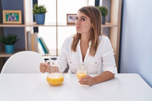 Young hispanic woman drinking glass of orange juice smiling looking to the side and staring away thinking. 