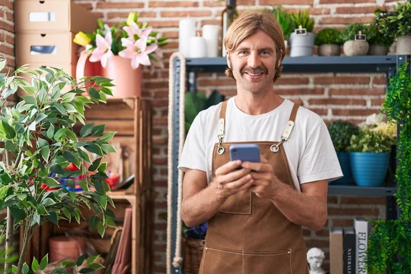 Young blond man florist smiling confident using smartphone at flower shop