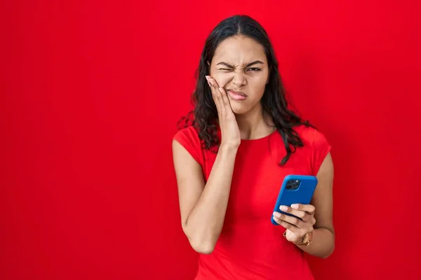 Młoda Brazylijka Używa Smartfona Czerwonym Tle Dotykając Ust Ręką Bolesnym — Zdjęcie stockowe