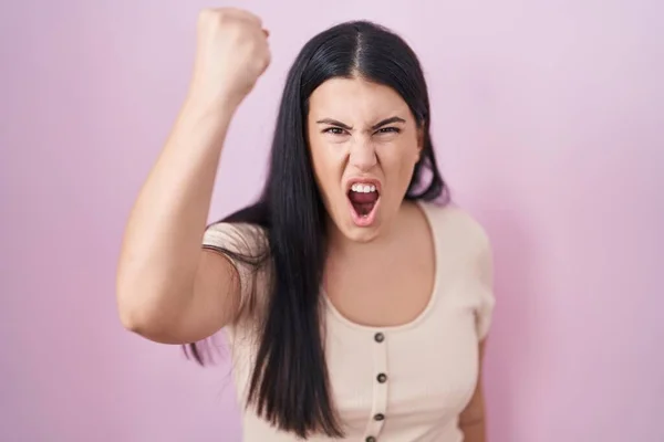 ピンク色の背景に立っている若いヒスパニック系の女性は怒りで叫んでいる間 怒りと怒りの拳を上げる怒っている 怒りと攻撃的な考え方 — ストック写真