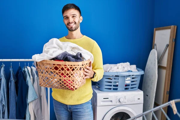 Hispanic man holding laundry basket smiling with a happy and cool smile on face. showing teeth.