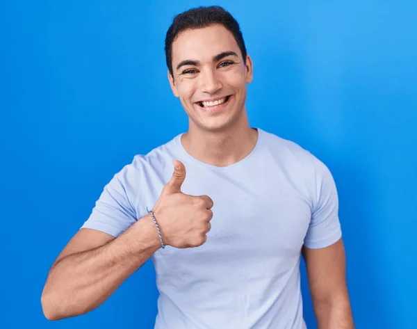青い背景の上に立つ若いヒスパニック系の男は手で幸せな親指をジェスチャーアップを行う 成功を示すカメラを見ると — ストック写真