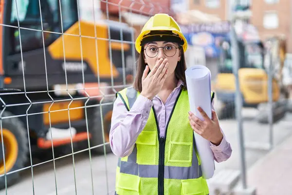 在建筑工地 西班牙裔女孩戴着建筑商的硬礼帽 用手捂住嘴 感到震惊和害怕犯错 惊讶的表情 — 图库照片
