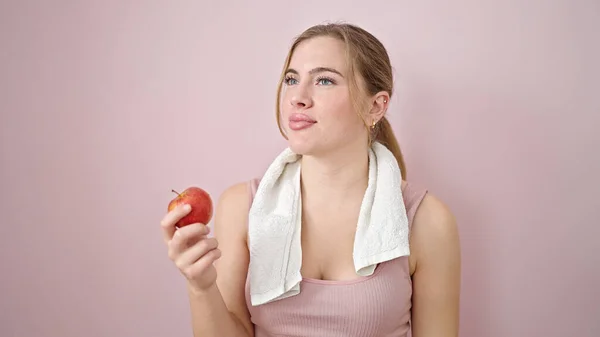 若いですブロンド女性身に着けていますスポーツウェア保持リンゴ上の隔離されたピンクの背景 — ストック写真