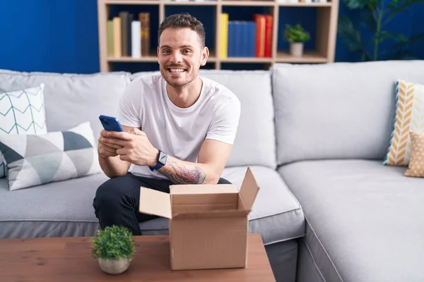 Young Hispanic Man Using Smartphone Unpacking Cardboard Box Home — Stock Photo, Image