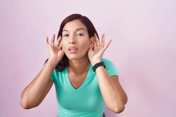 若いヒスパニック系の女性ピンクの背景に立って耳のジェスチャーで両方の手を聞こうとすると ゴシップへの好奇心 聴覚障害や聴覚障害 — ストック写真