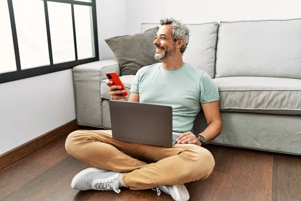 Stock image Middle age grey-haired man using smartphone and laptop sitting on floor at home