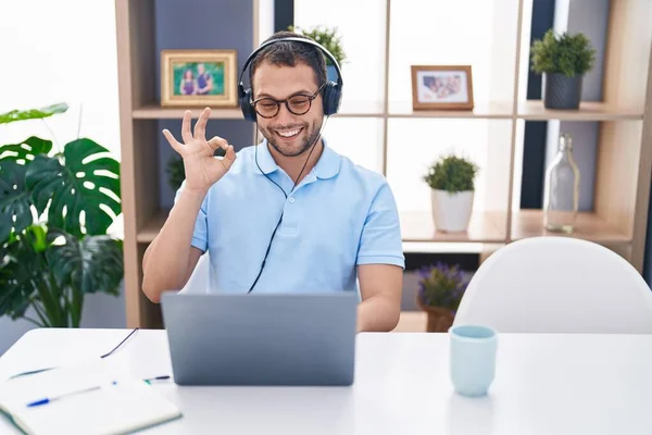 指でOkサインを行うヘッドフォンを身に着けているコンピュータのラップトップを使用して作業ヒスパニック系の男 笑顔優しいジェスチャー優れたシンボル — ストック写真