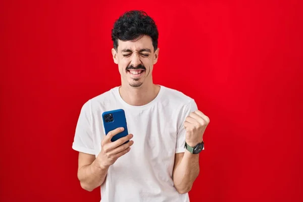 Hombre Hispano Usando Teléfono Inteligente Sobre Fondo Rojo Muy Feliz —  Fotos de Stock