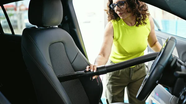 Middle age hispanic woman vacuuming car interior with vacuum cleaner at car wash station