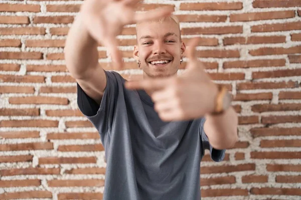 レンガの壁に立っている若い白人男性は 幸せな顔をした手と指でフレームを作って笑っています 創造性と写真の概念 — ストック写真