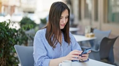 Akıllı telefon kullanan genç ve güzel bir İspanyol kadın kahve dükkanının terasında oturmuş gülümsüyor.