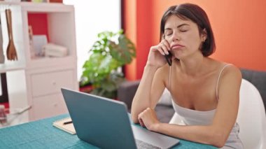 Genç, güzel, İspanyol bir kadın yemek odasında bilgisayarla akıllı telefondan konuşuyor.