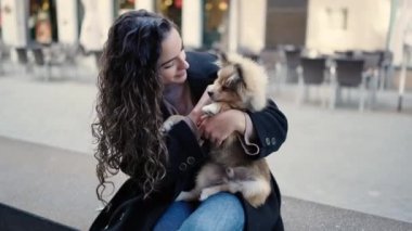 Köpeğiyle gülümseyen genç İspanyol kadın sokakta bankta oturuyor.