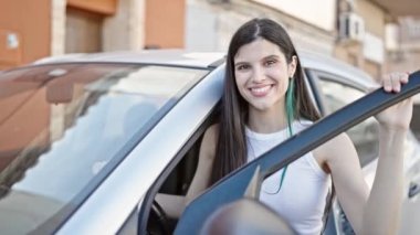 Genç, güzel, İspanyol bir kadın gülümsüyor. Sokakta araba kapısını açıyor.