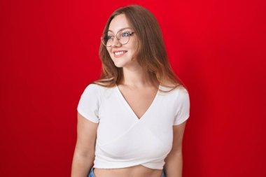 Kırmızı arka planda duran beyaz bir kadın yüzünde gülümseme, doğal bir ifade ile yan yana bakıyor. Kendinden emin gülen. 