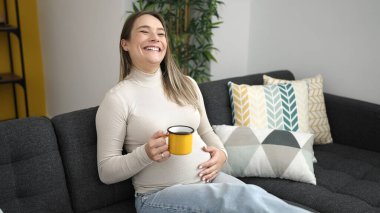 Young pregnant woman drinking coffee touching belly at home
