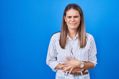 Hispanic young woman standing over blue background with hand on stomach because indigestion, painful illness feeling unwell. ache concept. 