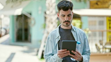 Latin kökenli genç bir adam, kahve dükkanının terasında elinde su şişesi olan rahat bir ifadeyle dokunmatik ped kullanıyor.