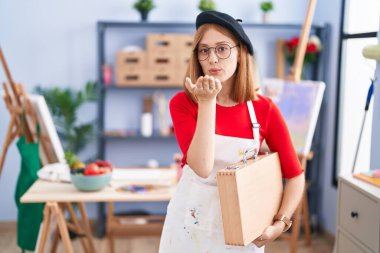 Sanat stüdyosundaki genç kızıl saçlı kadın elinde sanat çantası ile kameraya bakıyor ve havada bir öpücük savuruyor. Güzel ve seksi. aşk ifadesi. 