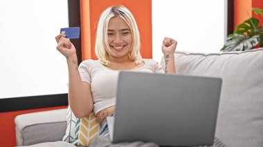 Laptop ve kredi kartıyla alışveriş yapan genç ve güzel bir İspanyol kadın evdeki kazanan ifadesiyle koltukta oturuyor.