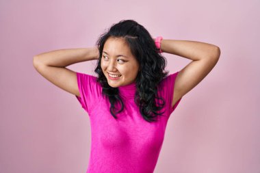 Young asian woman standing over pink background relaxing and stretching, arms and hands behind head and neck smiling happy 