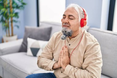 Orta yaşlı, kır saçlı, yoga egzersizi yapan, evdeki koltukta oturan bir adam.