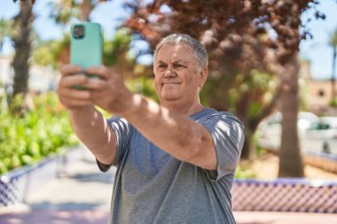 Orta yaşlı, gri saçlı bir adam parkta rahat bir ifadeyle akıllı telefondan selfie çekiyor.