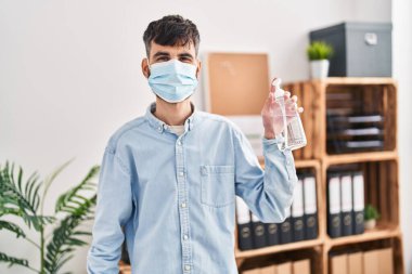 Young hispanic man business worker wearing medical mask using sanitizer gel hands at office