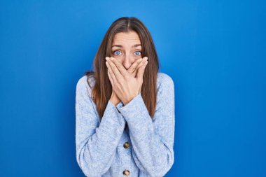 Young woman standing over blue background shocked covering mouth with hands for mistake. secret concept. 