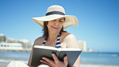 Orta yaşlı İspanyol bir turist havluda güneşleniyor sahilde kitap okuyor.