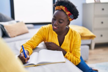 Genç Afrikalı Amerikalı kadın deftere yazı yazıyor. Evinde yerde oturuyor.