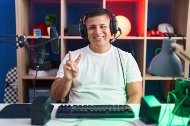 Young hispanic man playing video games showing and pointing up with fingers number two while smiling confident and happy. 