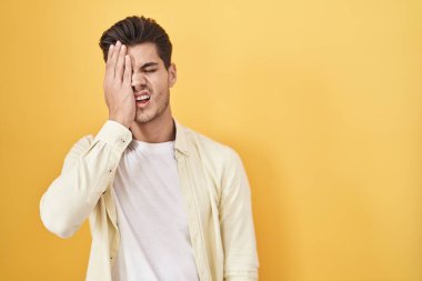 Sarı arka planda dikilen İspanyol genç adam yarı yüzünü, göz ve ağzını eliyle kapatmaktan yoruldu. Yüzüm acıyor.. 