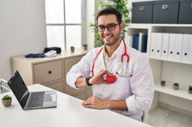 Young hispanic man wearing doctor uniform holding heart over chest at clinic