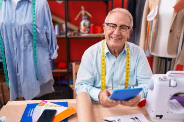 Orta yaşlı, gri saçlı terzi atölyedeki dokunmatik pedi kullanarak gülümsüyor.